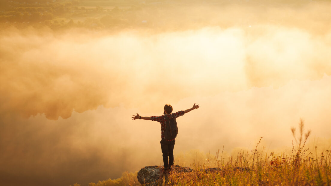 Anonymous male hiker standing on mountain slope with outstretched arms at sunrise