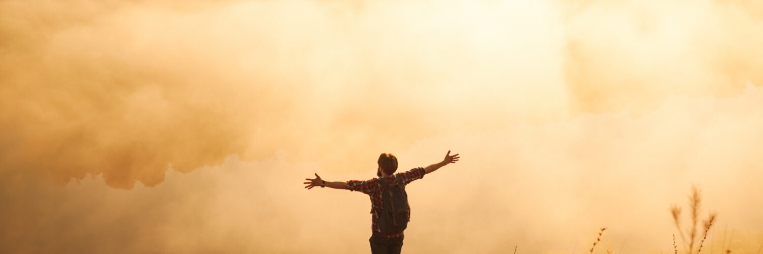Anonymous male hiker standing on mountain slope with outstretched arms at sunrise